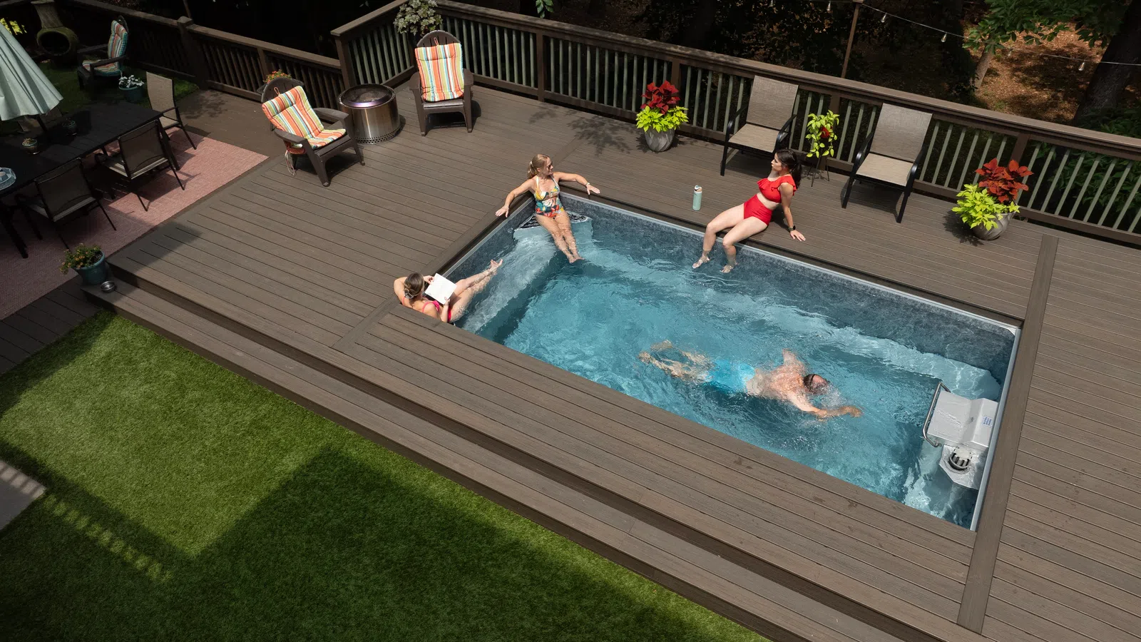 Aerial view of four people relaxing and swimming in a small pool on a wooden deck with colorful chairs and plants.