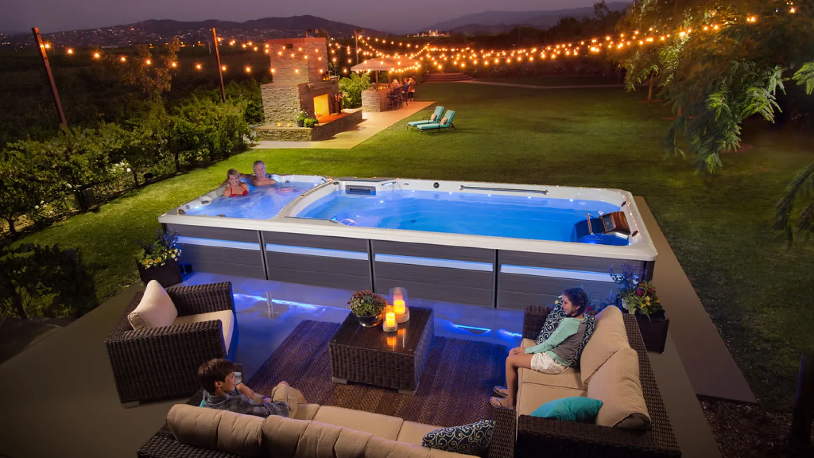 Evening backyard scene with people relaxing in a hot tub and on outdoor wicker furniture under string lights.