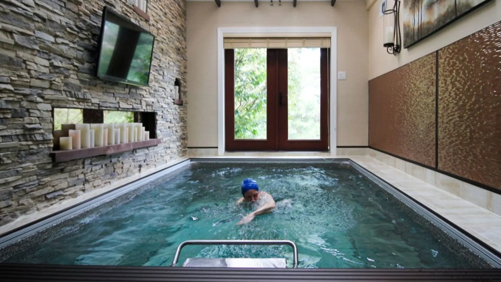 Indoor swim spa with swimmer, stone fireplace wall, candles, mounted TV, and glass doors leading outside.