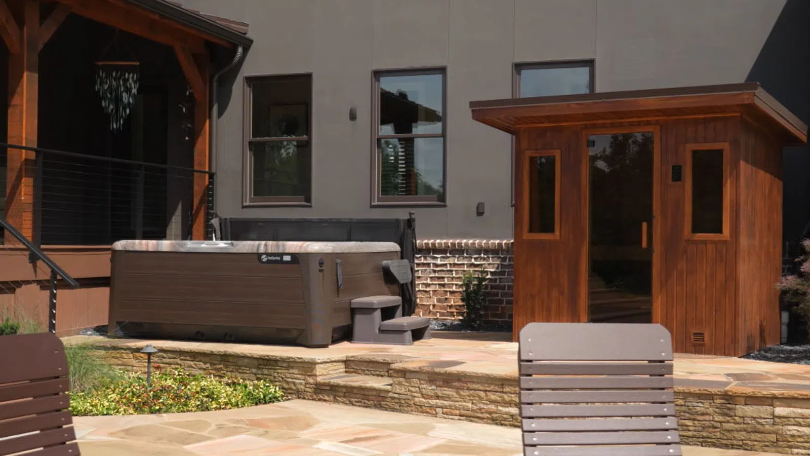 Modern backyard patio with hot tub, wooden sauna, and lounge chairs on stone flooring near house.