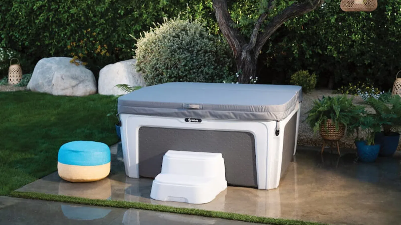Covered hot tub with white steps and blue stool in a garden surrounded by greenery and stone accents.