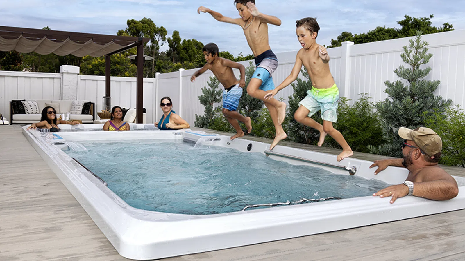 Three boys jump into a backyard swim spa while adults relax in and around the pool on a sunny day.