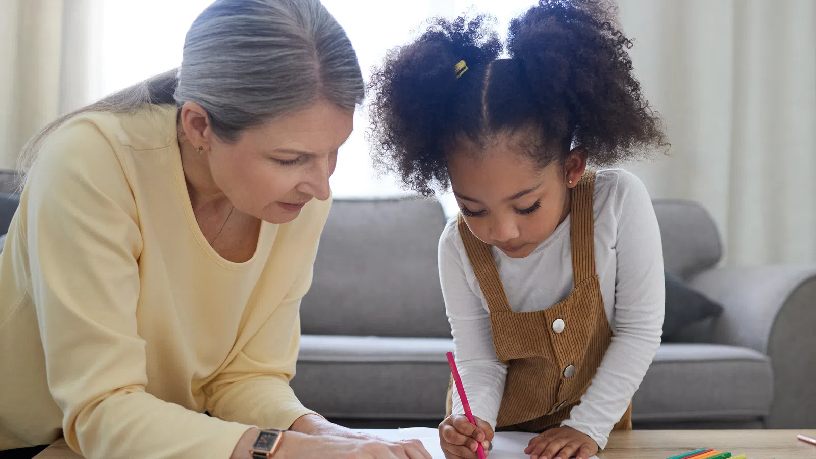 Young girl drawing with colored pencils while an older woman watches and guides her at a wooden table.
