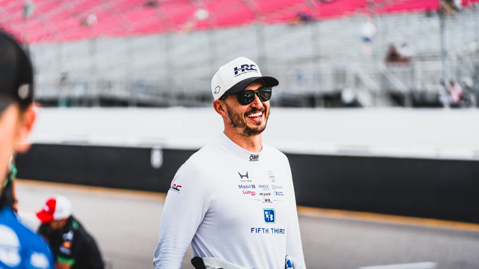 Smiling male race car driver wearing sunglasses and a white racing suit with sponsor logos in a racetrack pit area.