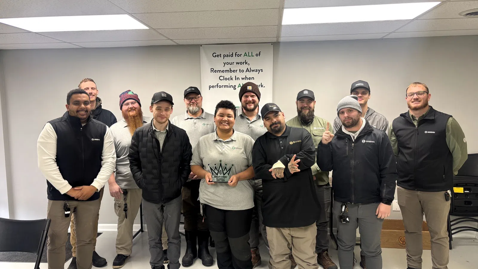 Group of twelve diverse workers in casual uniforms posing with awards in a break room with motivational poster.