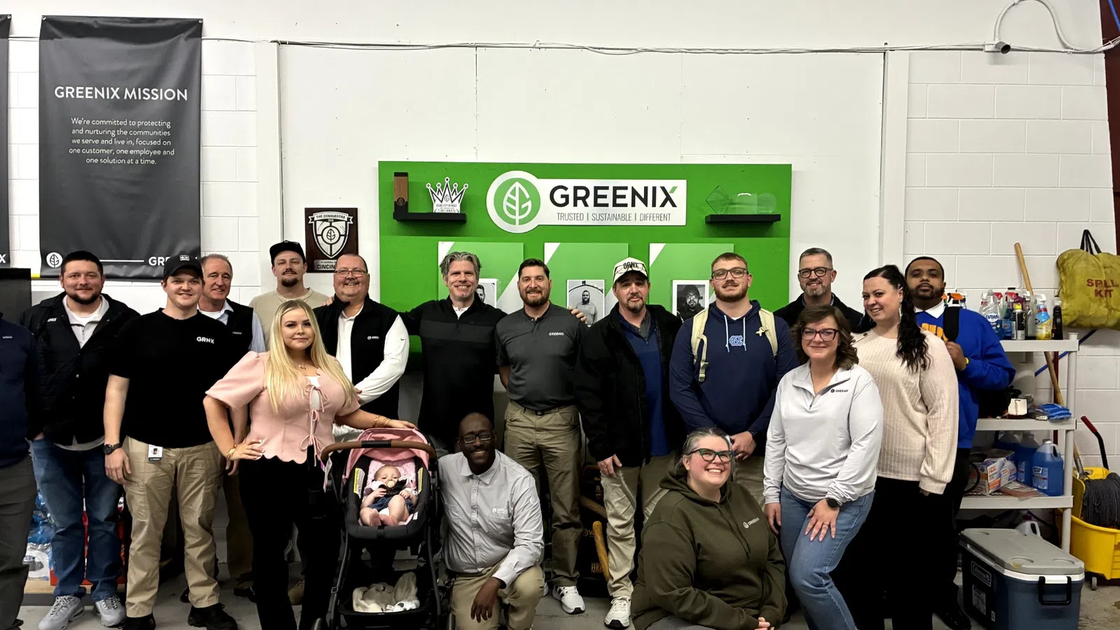 Group of diverse Greenix employees posing in front of Greenix mission and logo wall inside warehouse.