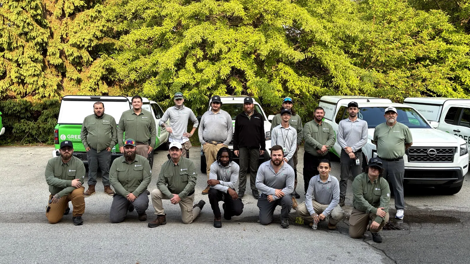 Group of landscaping workers posing in front of work trucks with green trees in the background outdoors.