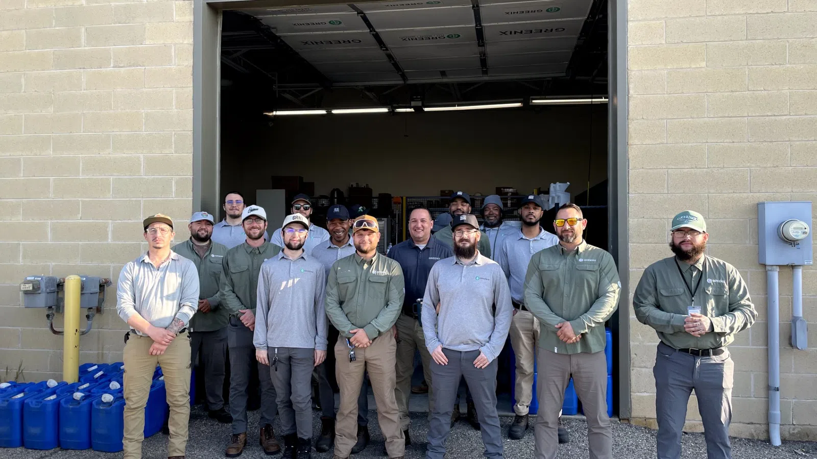 Group of fifteen men standing in front of an open warehouse door wearing casual work attire and caps.