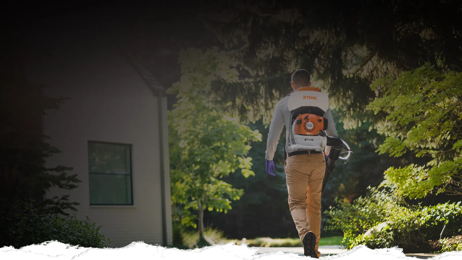 Person wearing a backpack leaf blower walking along a garden path near a house surrounded by green trees.