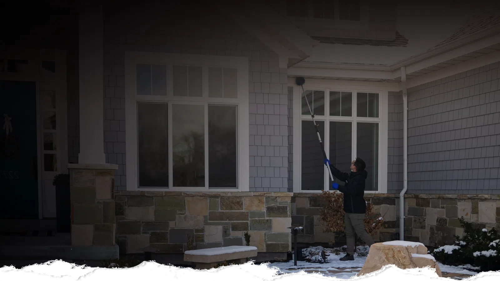 Person cleaning windows of a stone and shingles house during winter with snow on the ground