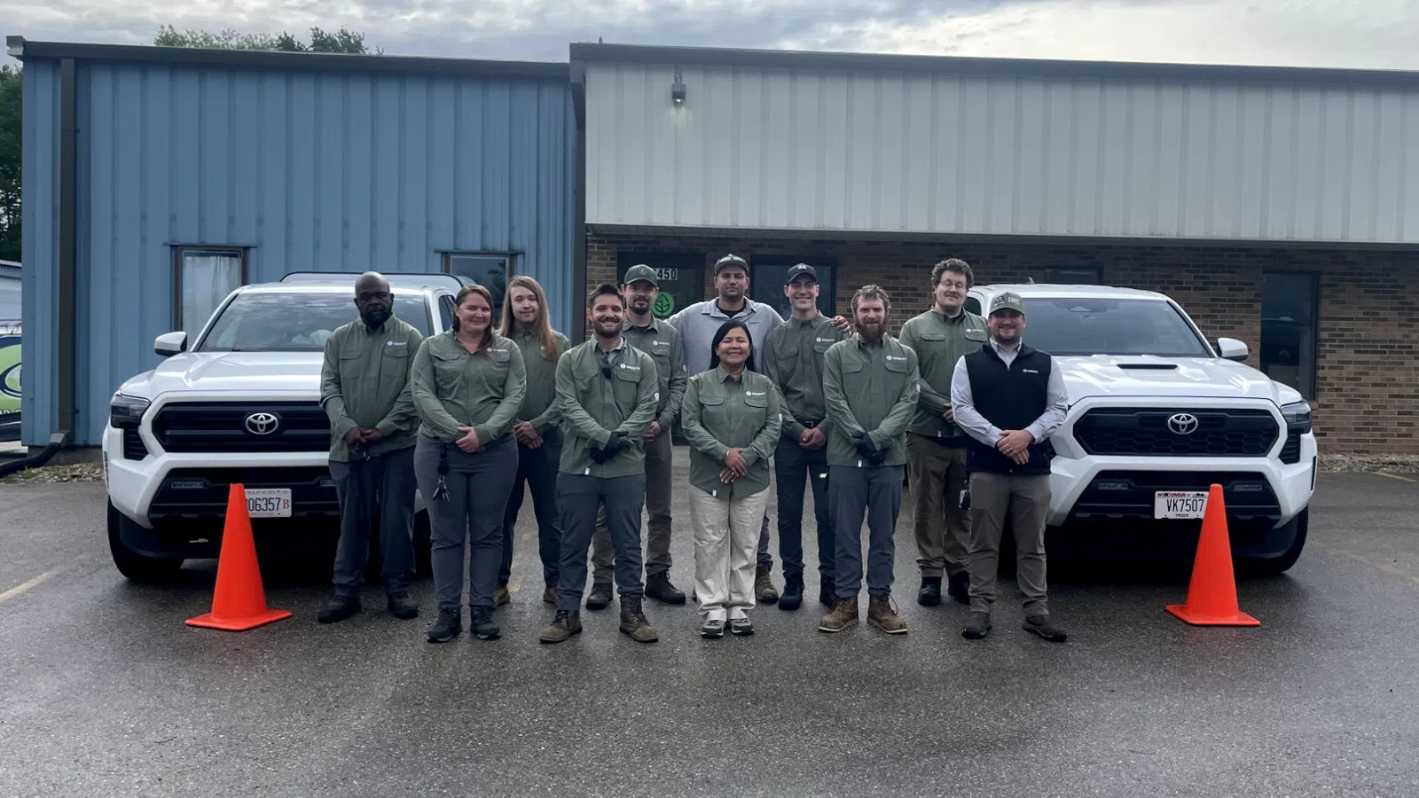 Group of eleven people standing in front of two white Toyota trucks outside a building on a cloudy day