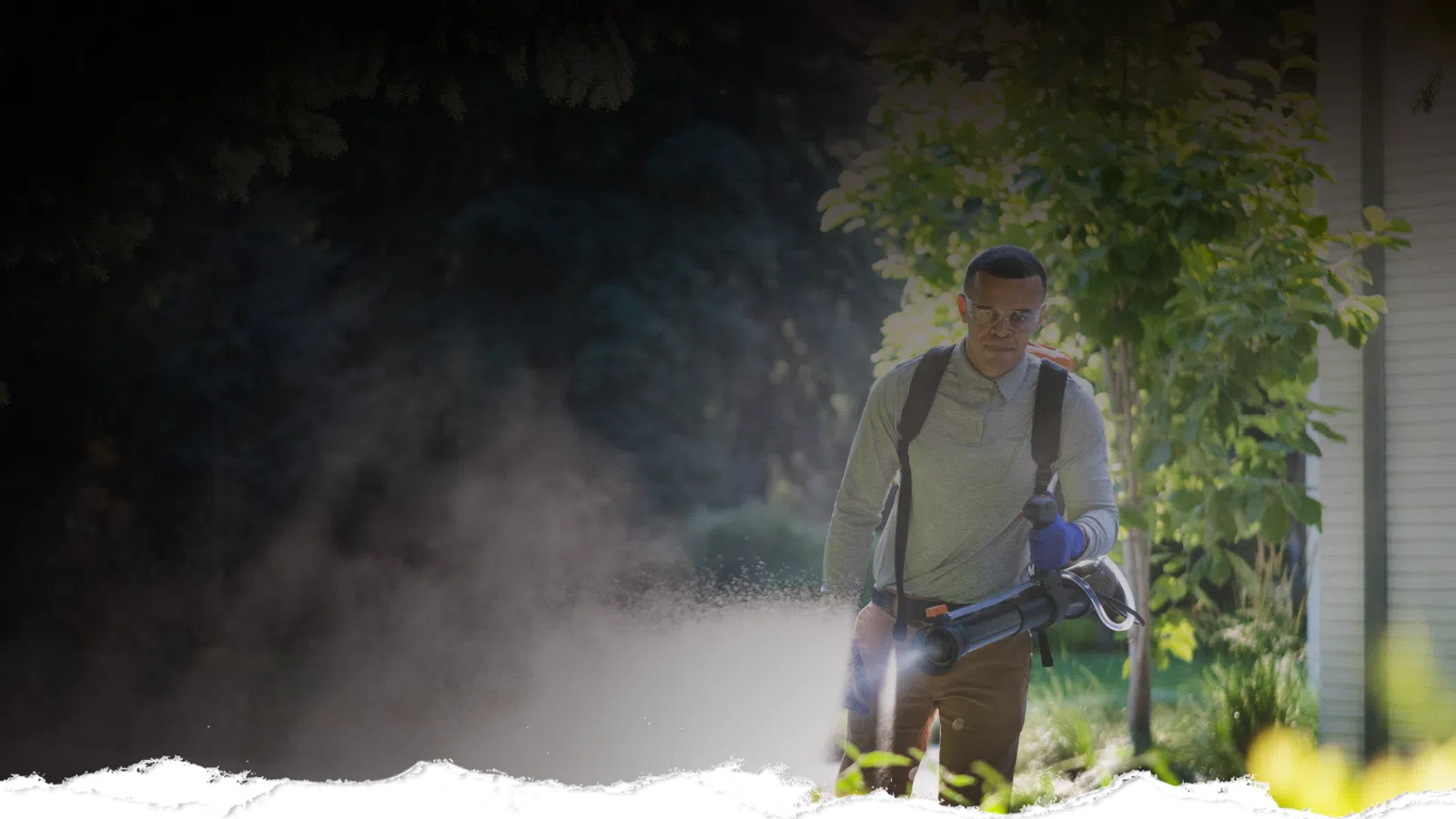 Man wearing protective gear using a fogger to spray pesticide outdoors near trees and a building.