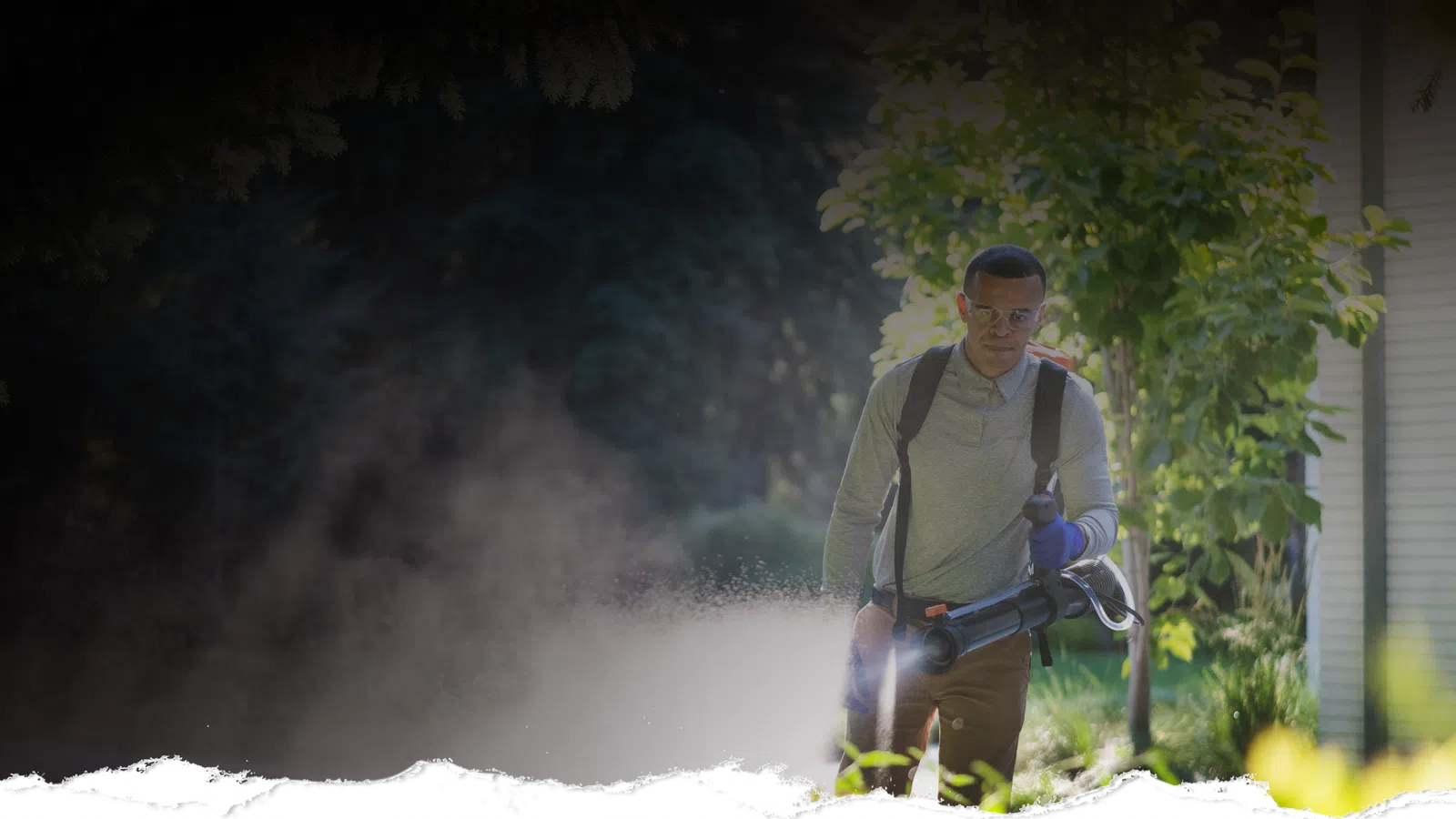 Man wearing backpack sprayer treating plants near a home’s stone and stucco exterior at dusk.