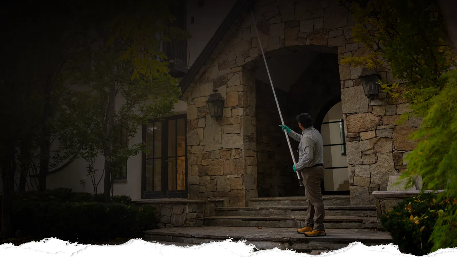 Man cleaning outdoor stone wall of a large house using a long pole tool near entrance steps.