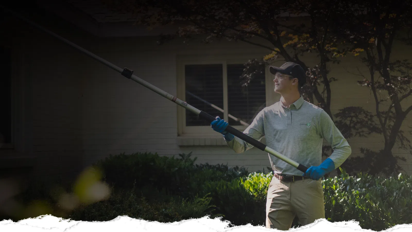 Man in gloves using a long pole tool to prune trees outside a house on a sunny day