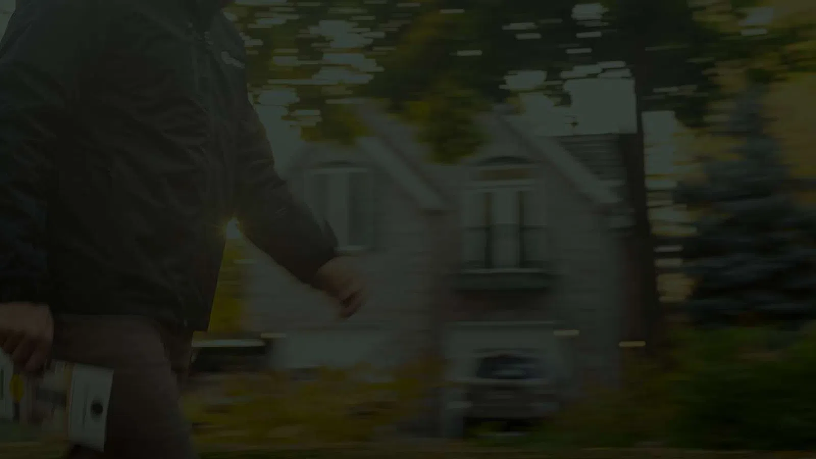 Person walking outside near houses on a tree-lined street holding a skateboard in one hand.