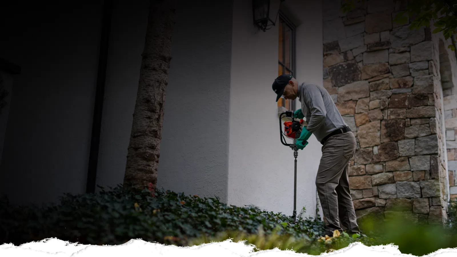 Man using a leaf blower to clear garden plants outside a stone and stucco house in daylight.