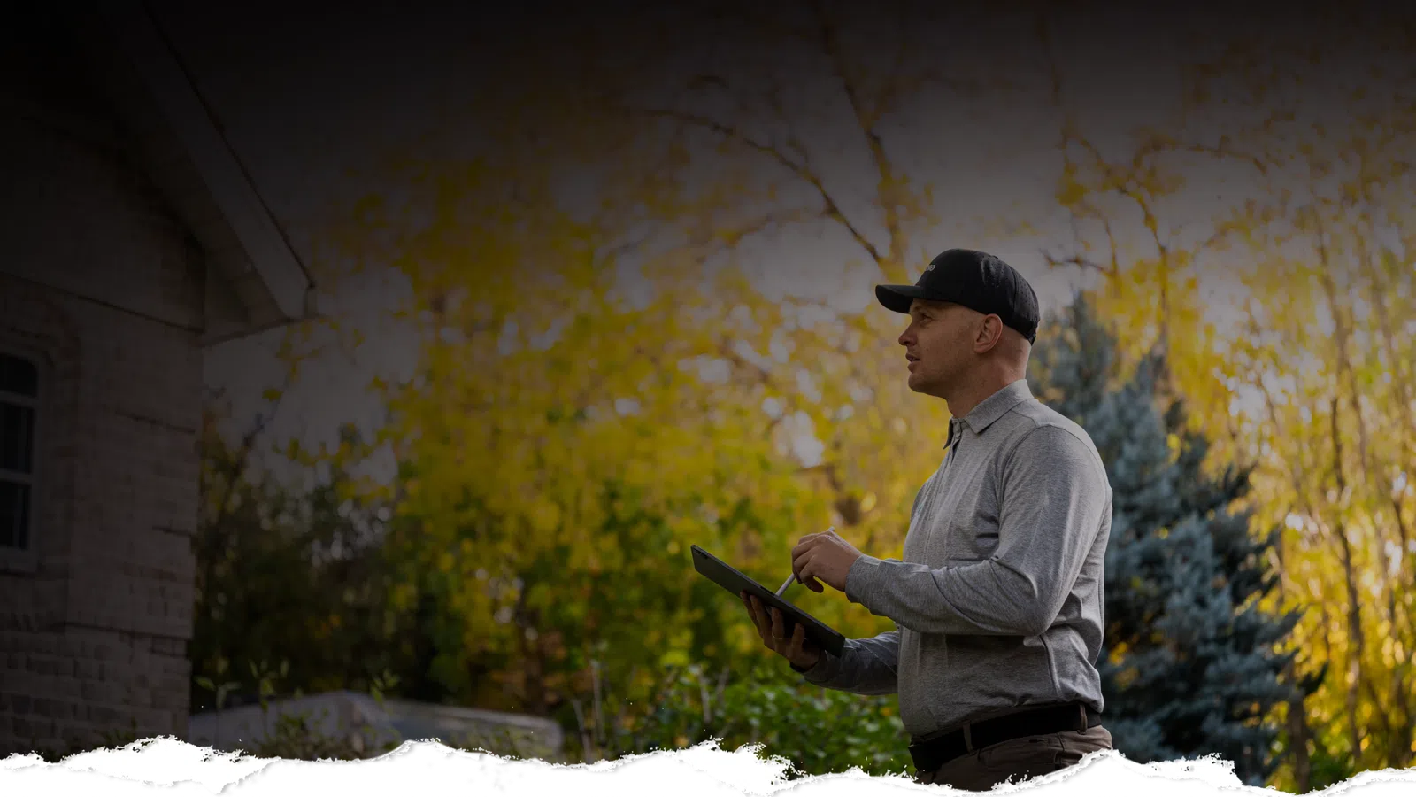 Home inspector wearing a cap holds a tablet outside a house surrounded by autumn trees and greenery.