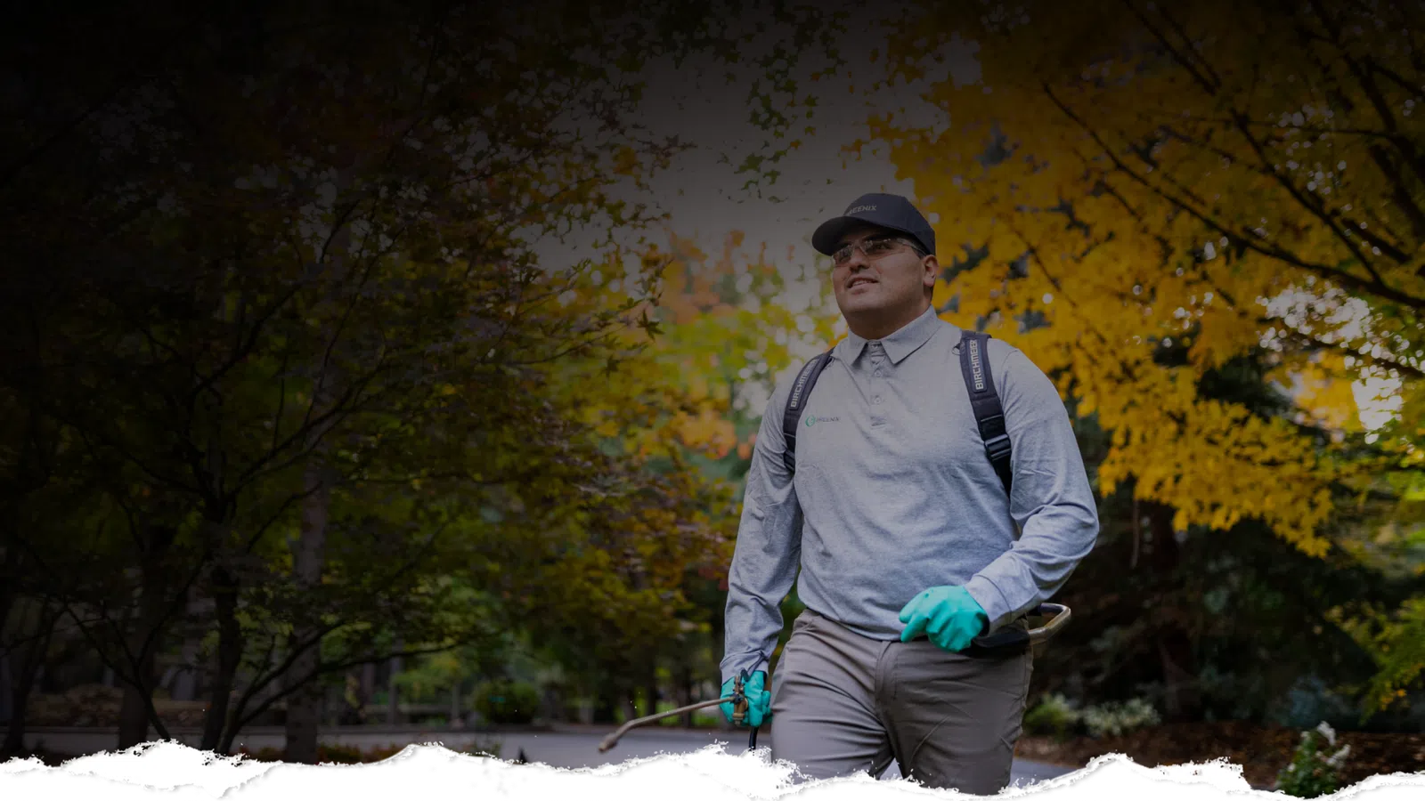 Man wearing gloves and backpack sprayer walking through a park with autumn foliage in the background