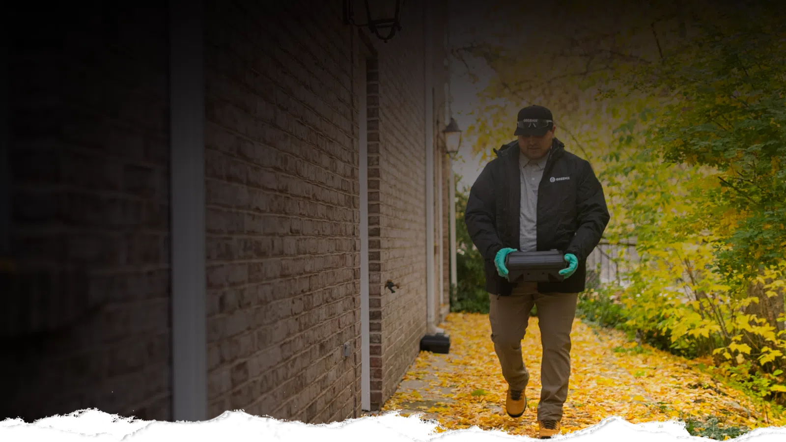 Delivery person walking on a leaf-covered path beside a brick building carrying a package wearing gloves and a jacket