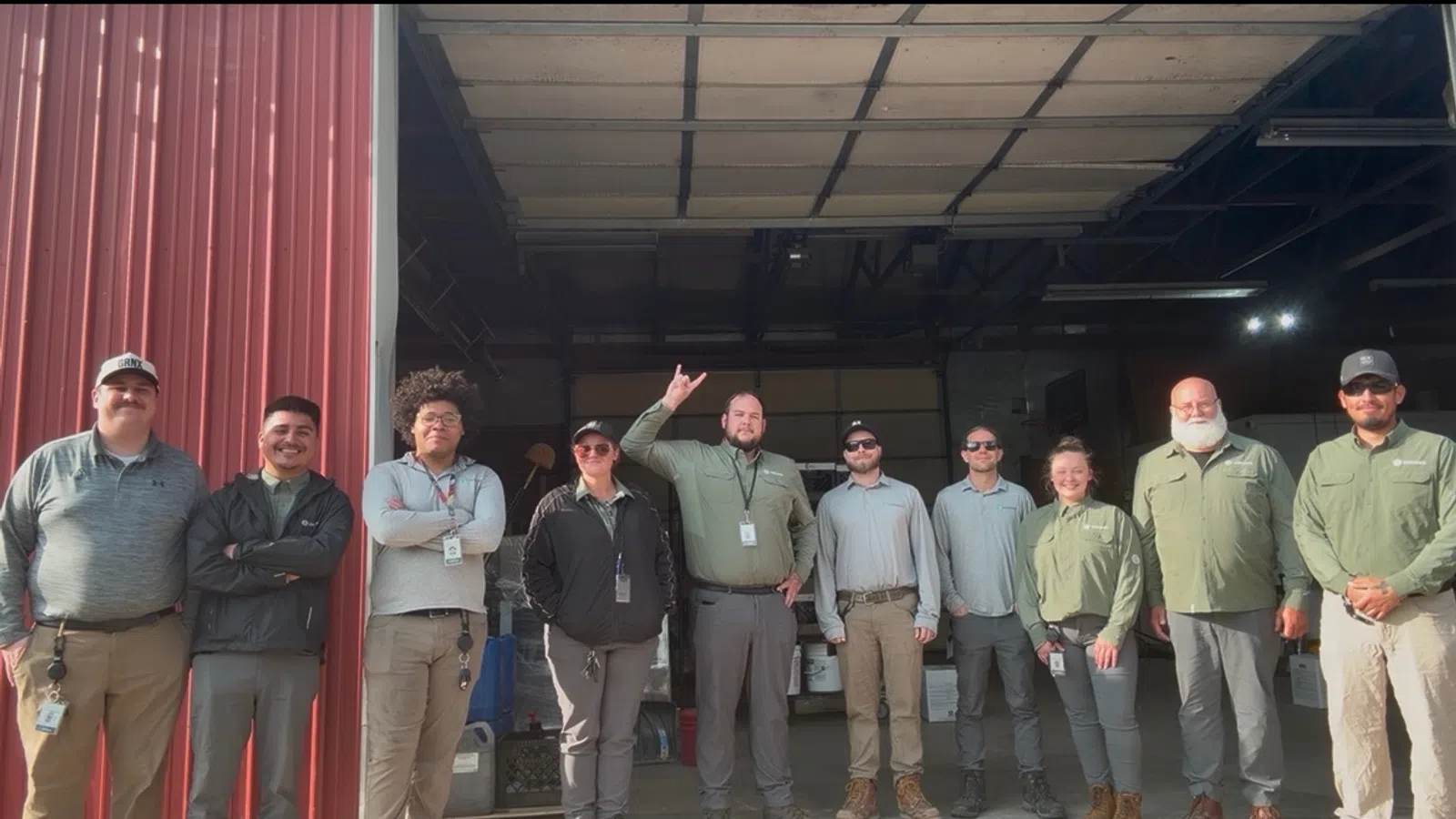 A group of ten diverse workers standing together outside a red building garage door, smiling under daylight.
