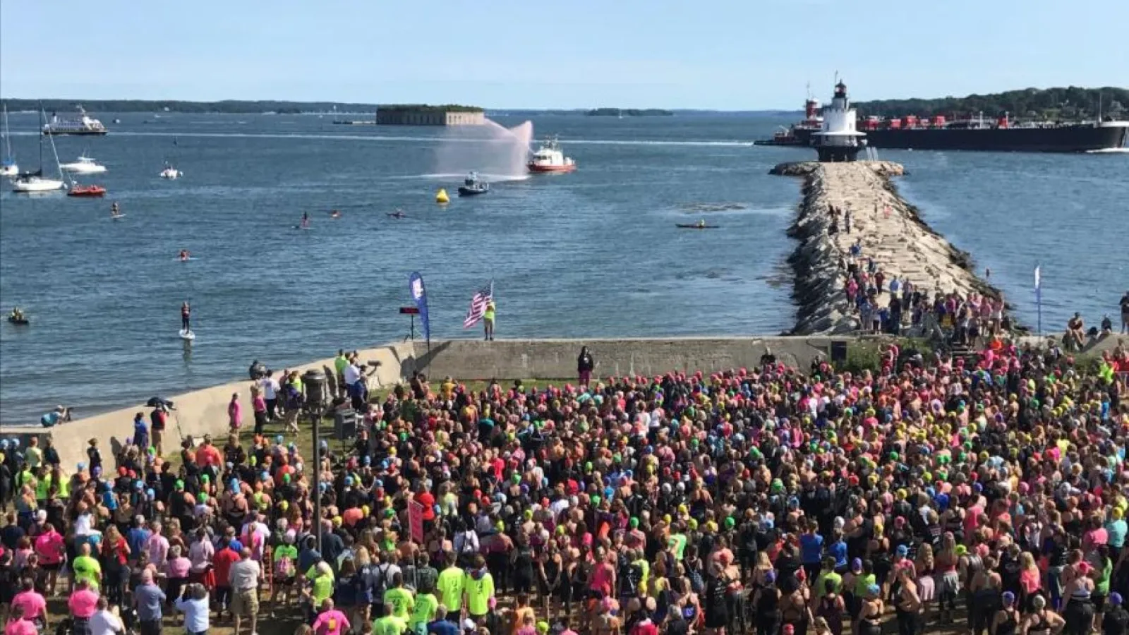 Large crowd gathered near the waterfront by a lighthouse on a sunny day with boats on the water.