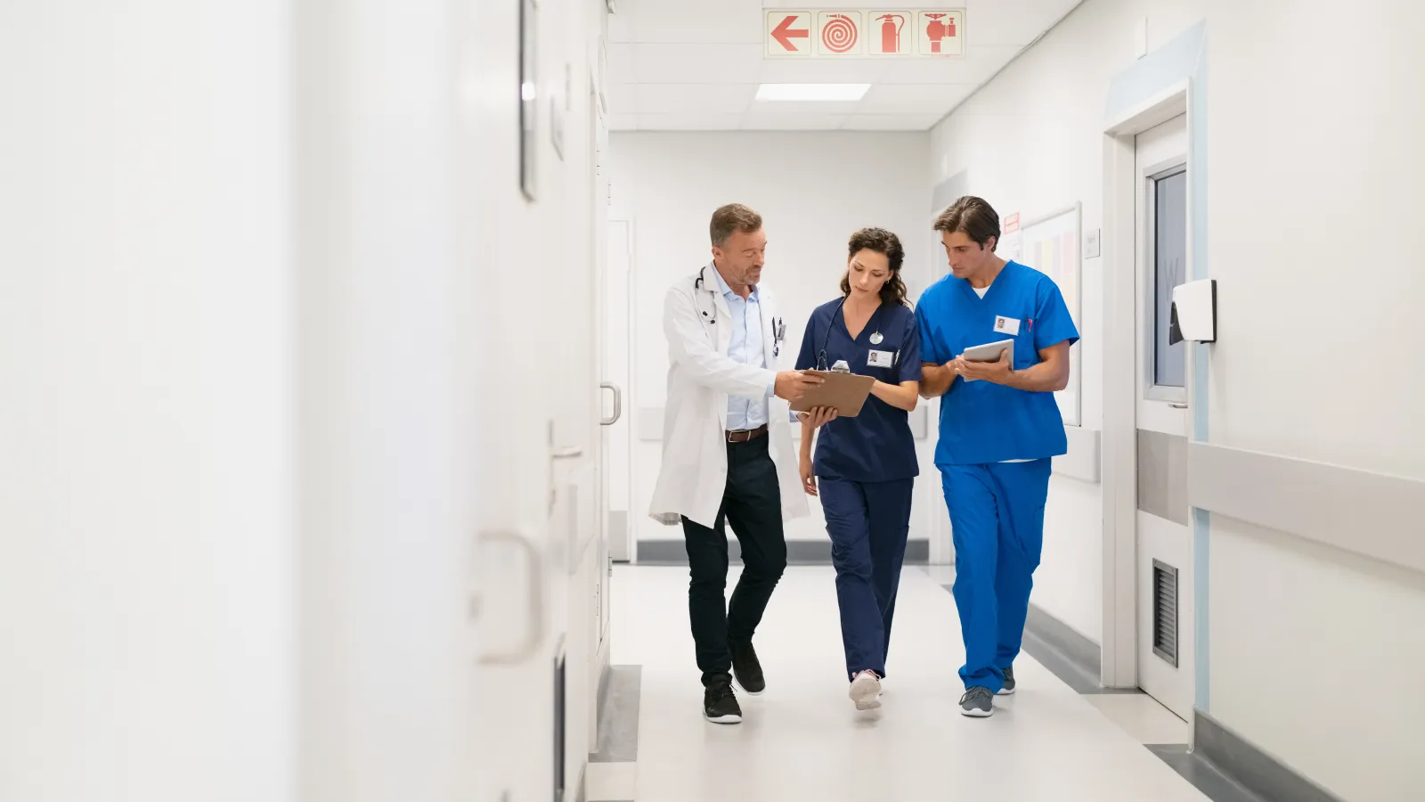Three healthcare professionals in scrubs and lab coat walking a hospital corridor discussing patient notes.