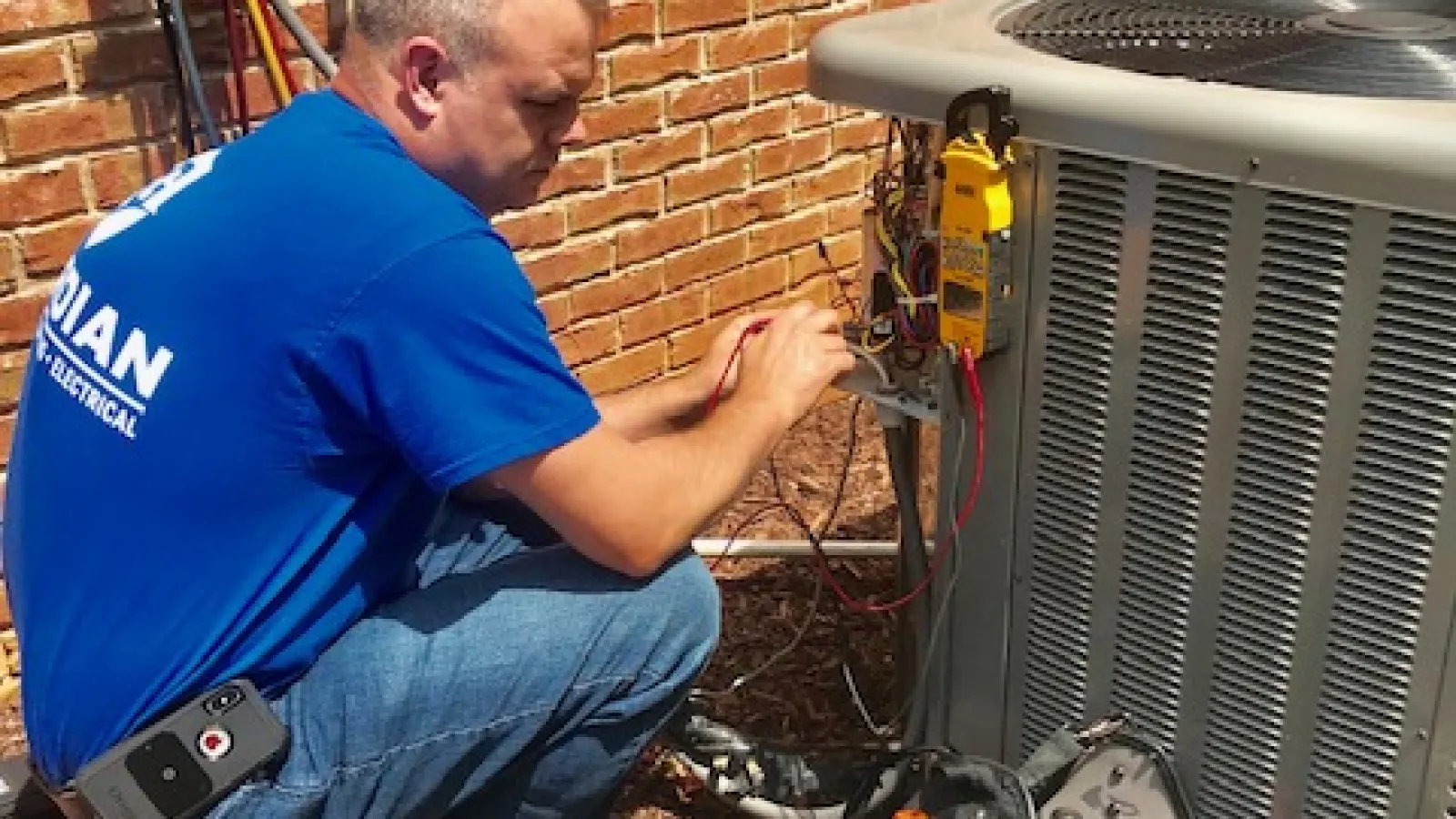Technician in blue shirt repairing an outdoor HVAC unit next to a brick wall with tools nearby