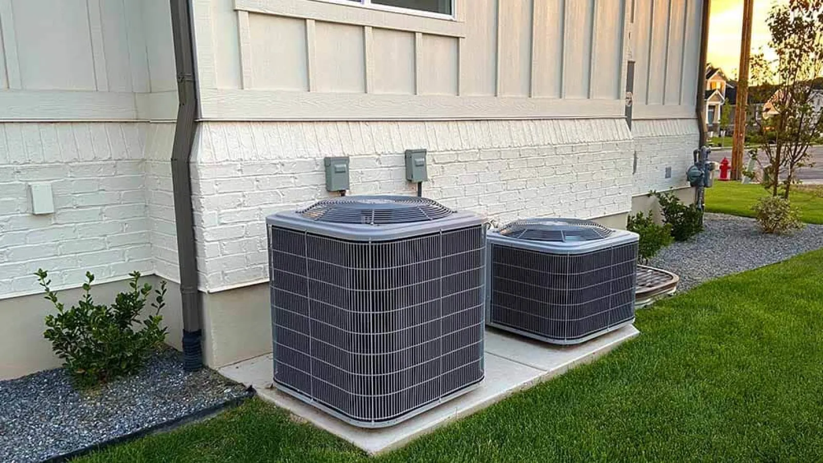 Two outdoor air conditioning units installed on concrete pads beside a white brick house with green lawn