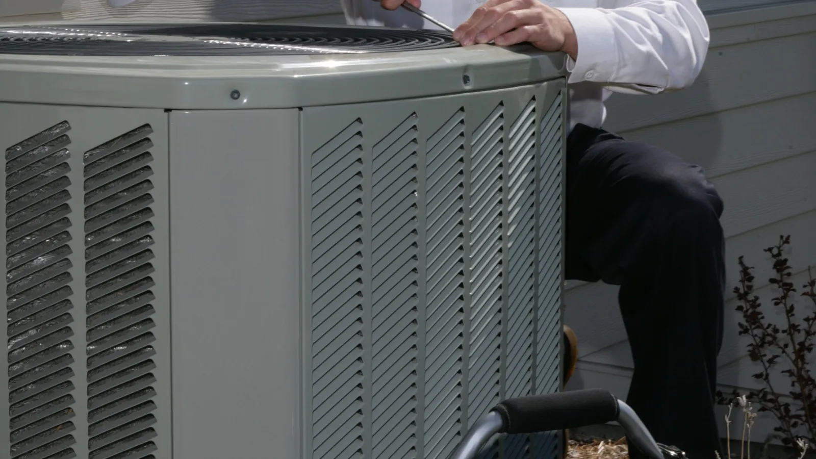 Technician repairing an outdoor air conditioning unit with tools in a black tool bag nearby beside a house.