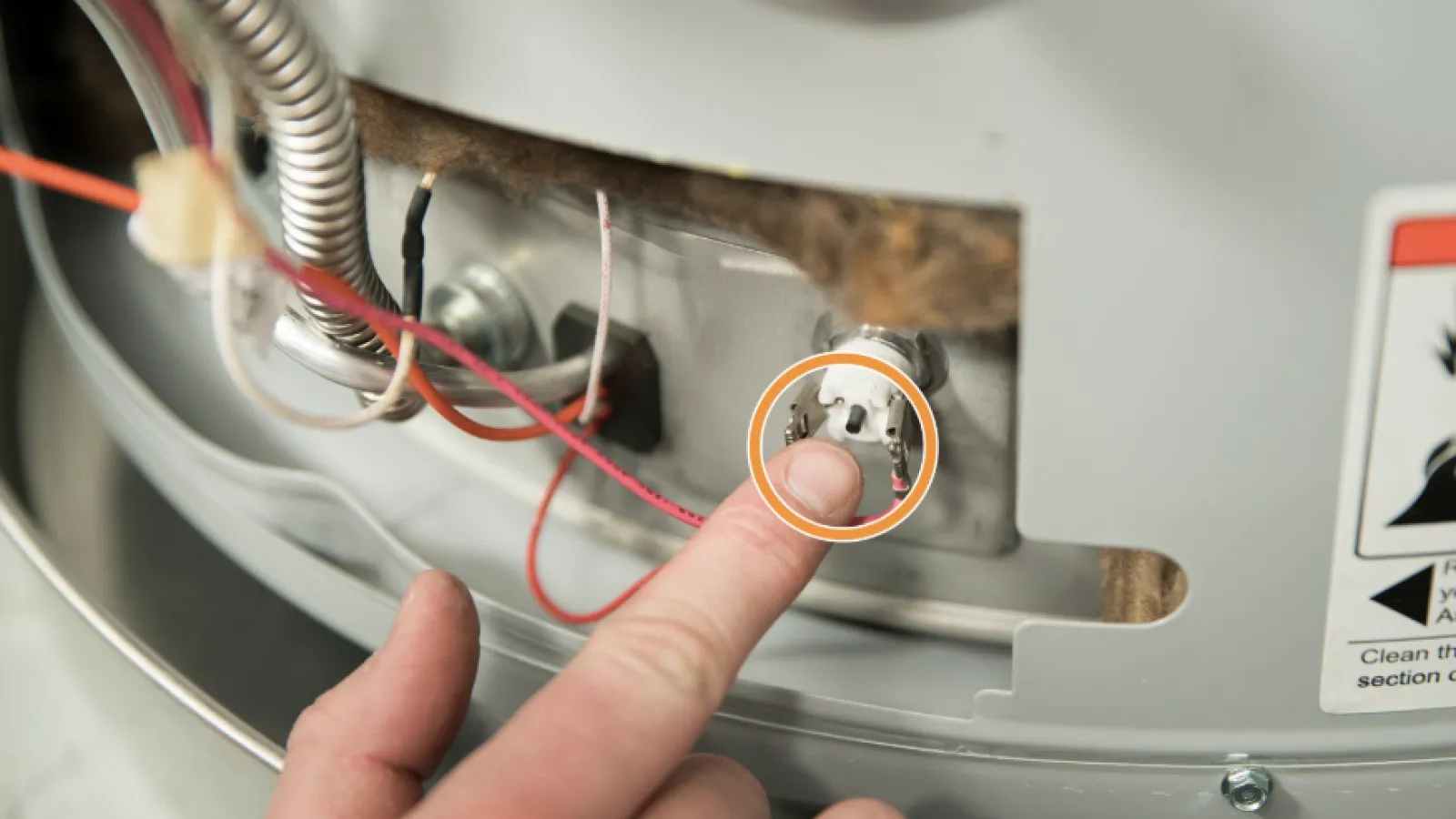 Person's finger pressing a reset button on a water heater with wires and metal components visible.