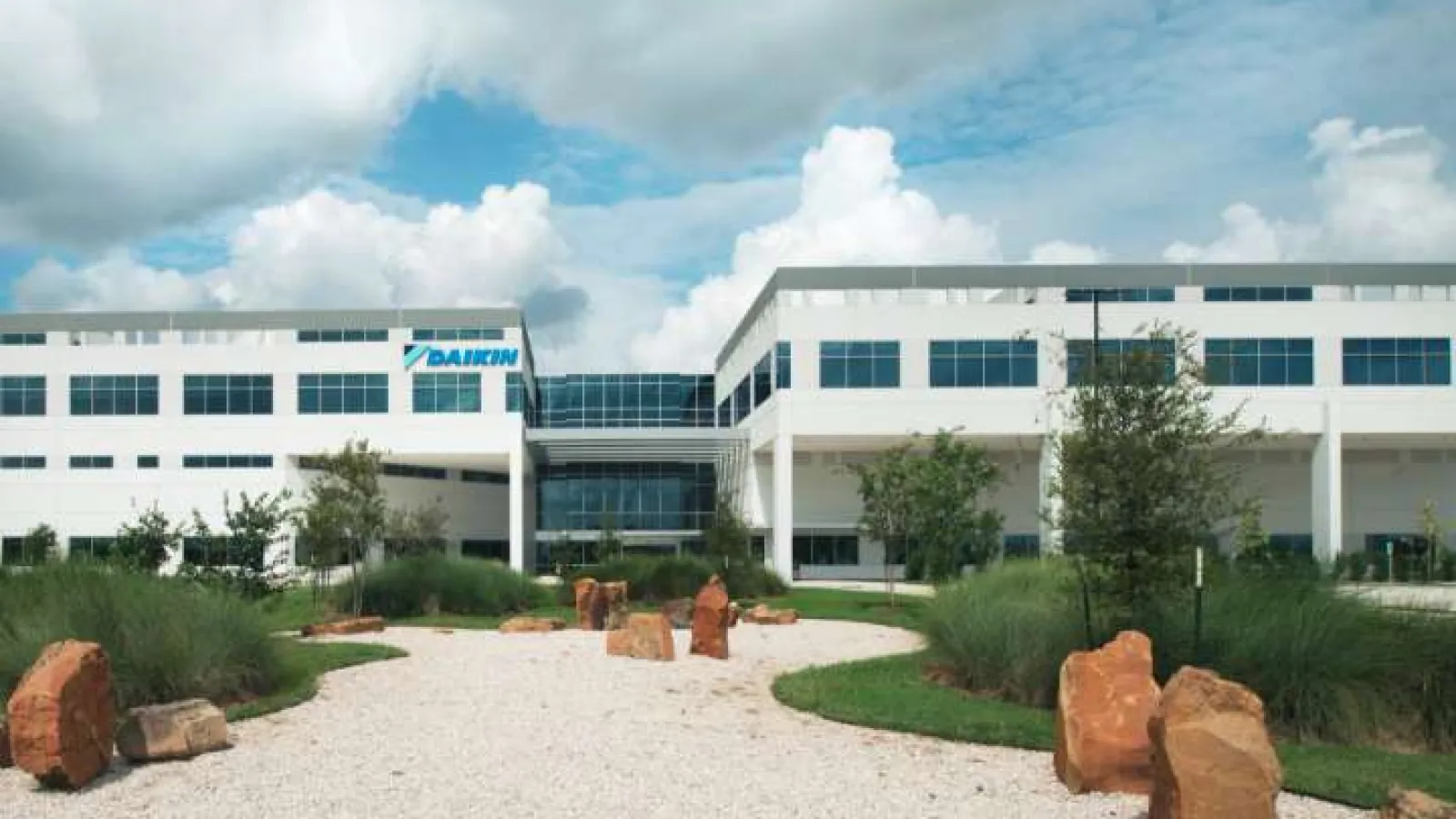 Modern white office building surrounded by landscaped greenery, rocks, and a gravel pathway under blue sky.