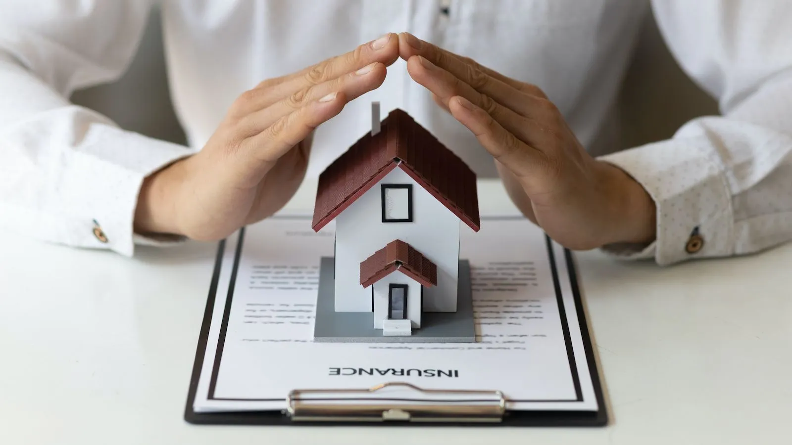 Hands protect a miniature house model over an insurance policy document on a clipboard.