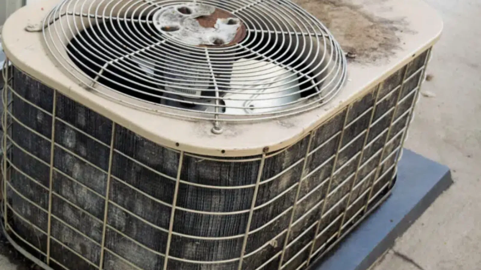 A weathered air conditioning unit showing dirt and wear, sitting on a concrete base outdoors.