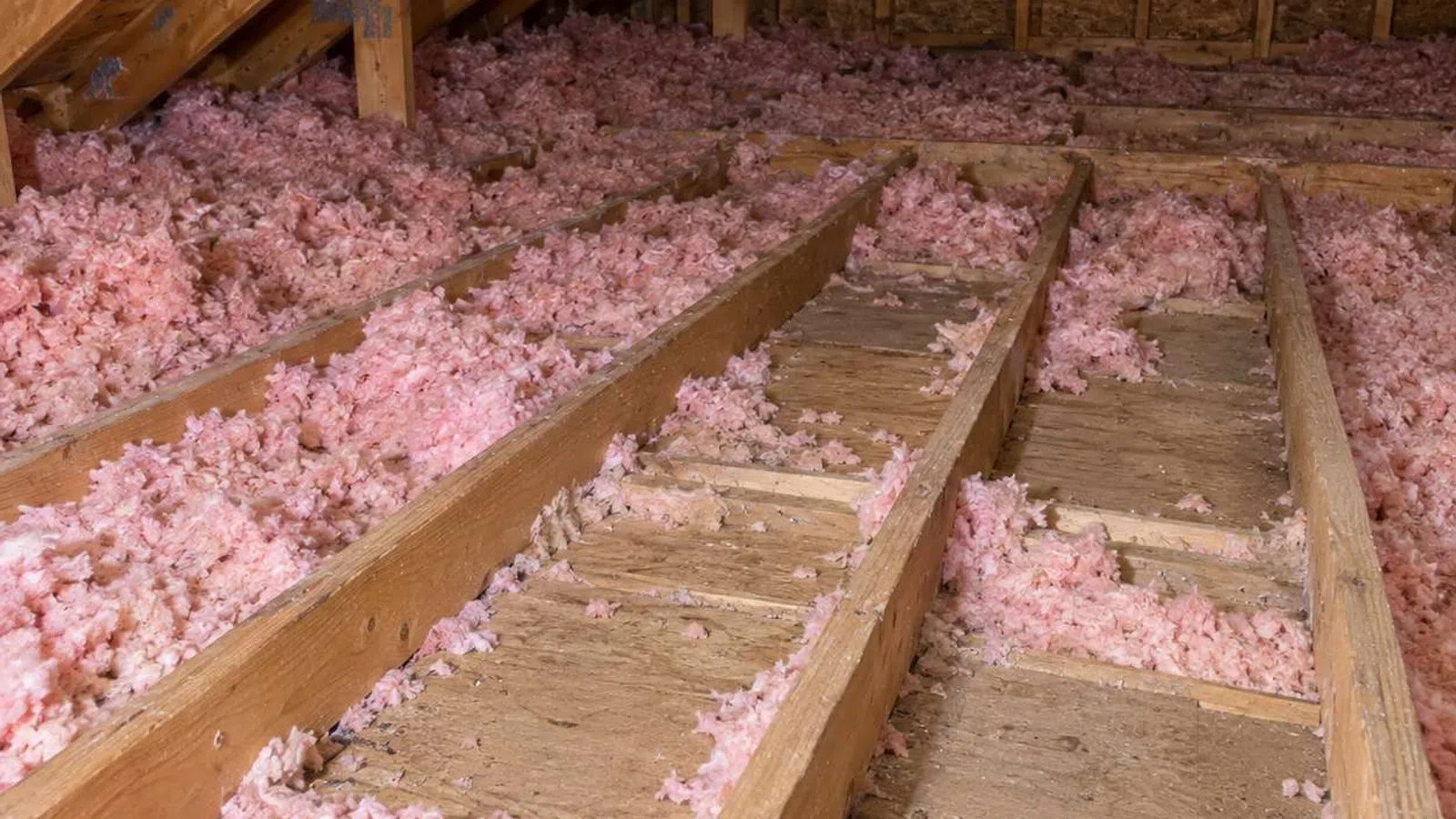 Attic space with exposed wooden beams and pink fiberglass insulation between floor joists under roof.