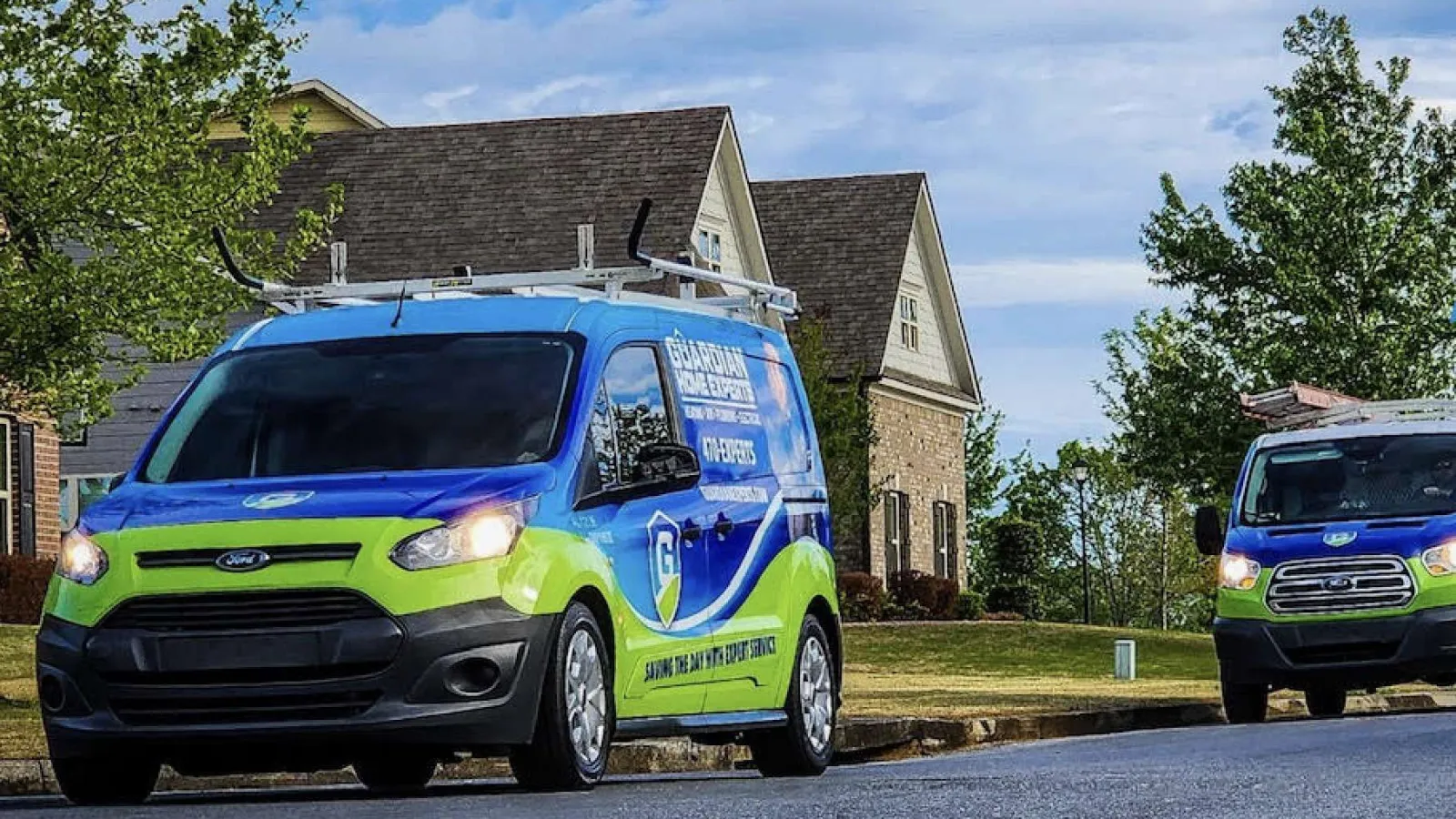 Two blue and green service vans parked on a suburban street near houses with trees and a partly cloudy sky.