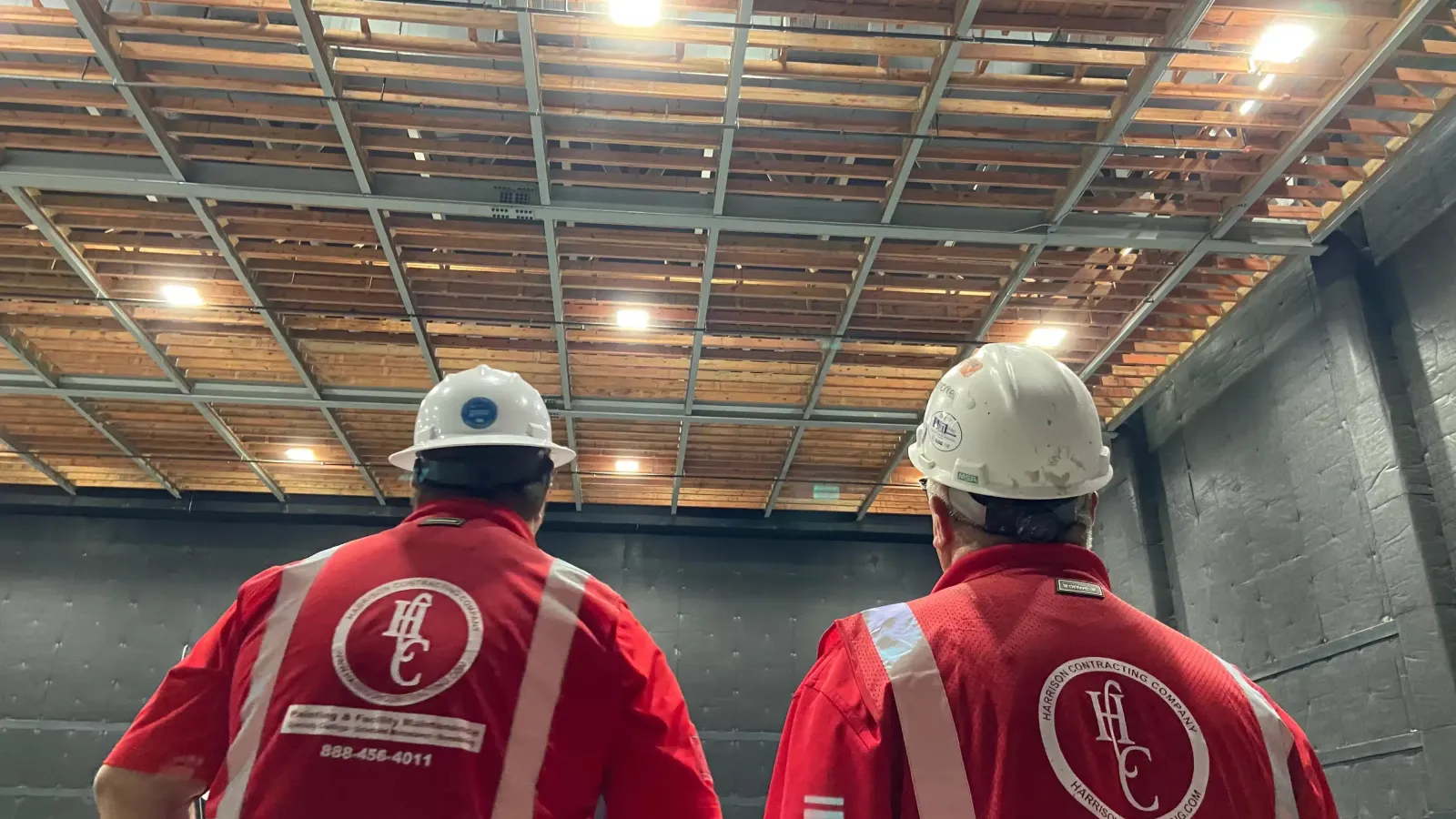 Two construction workers in red shirts and white helmets inspecting a warehouse ceiling with lights and exposed beams.