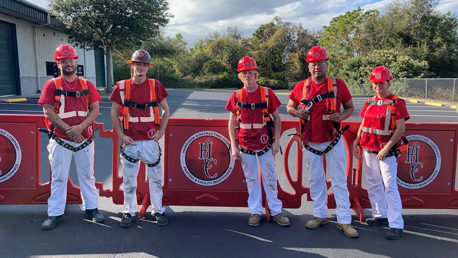 Five construction workers wearing red safety helmets and harnesses standing in front of red barriers with company logo outdoors.