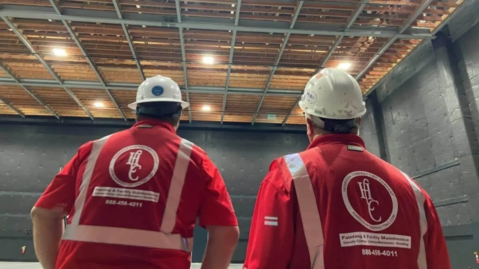 Two construction workers in red safety vests and white helmets inspecting an unfinished indoor ceiling.