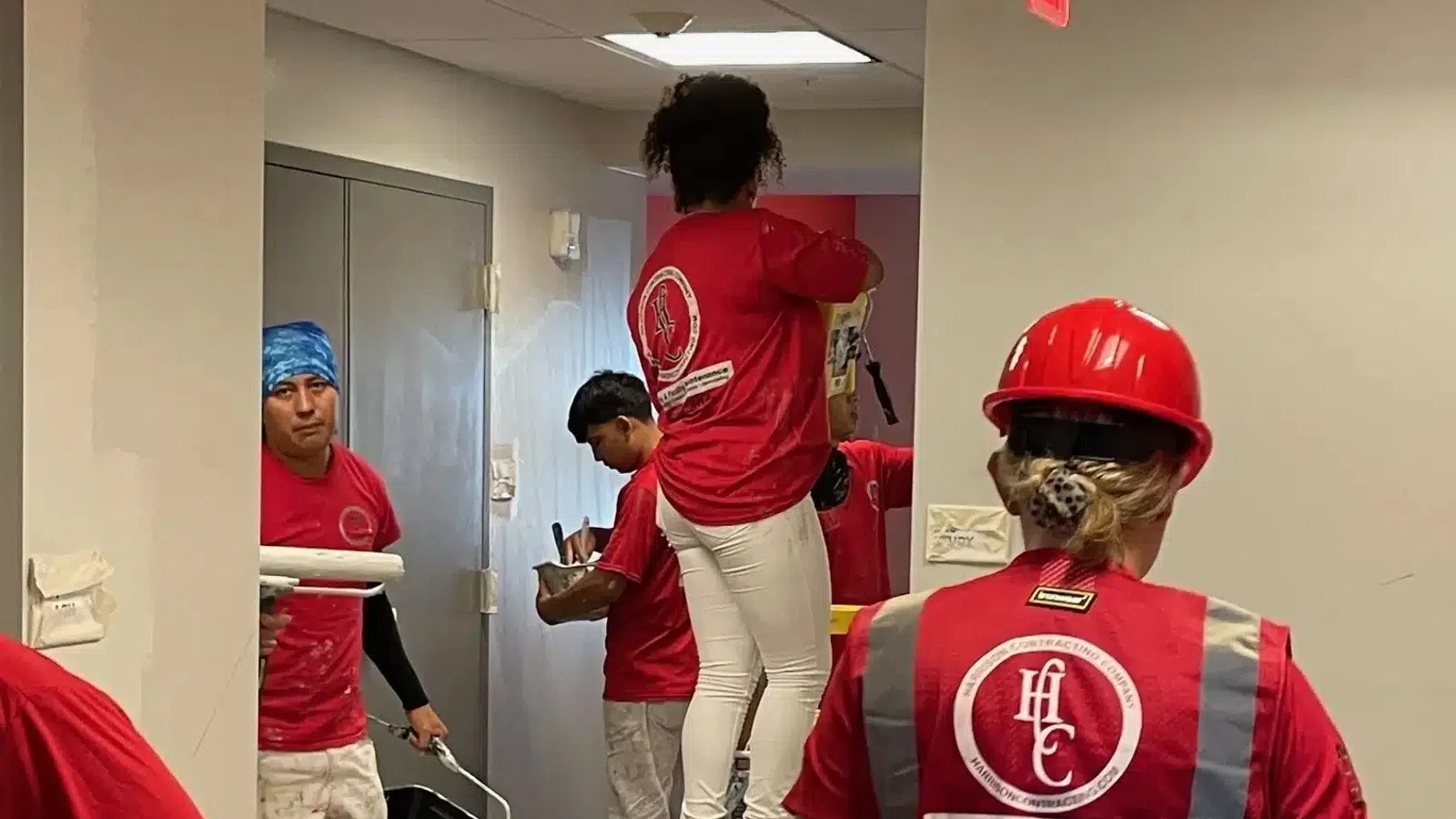 Group of painters in red shirts working indoors, some painting walls and others using tools inside a building hallway.
