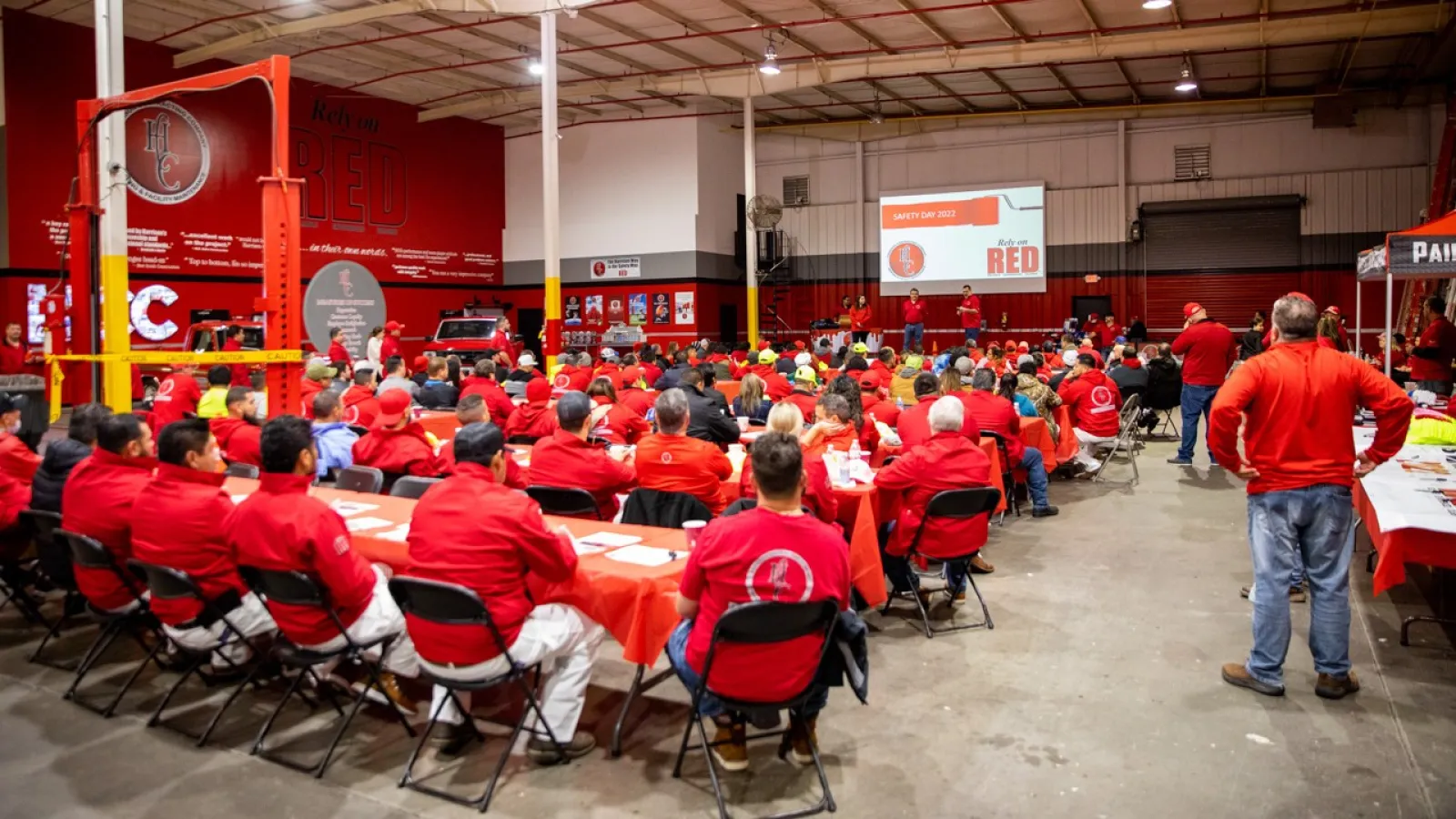 Large group of people wearing red gathered in a warehouse for a safety day presentation with a projector screen.