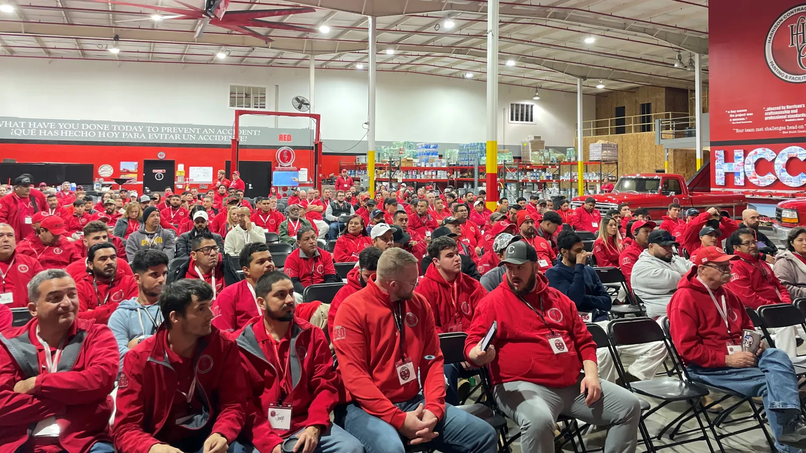 Large group of people in red jackets seated indoors attending a safety or training event in a warehouse setting.