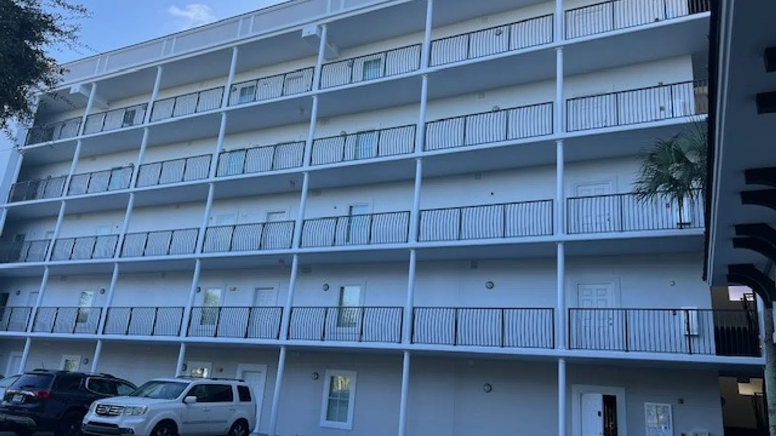 Four-story white apartment building with exterior balconies and parked cars in foreground under clear sky.