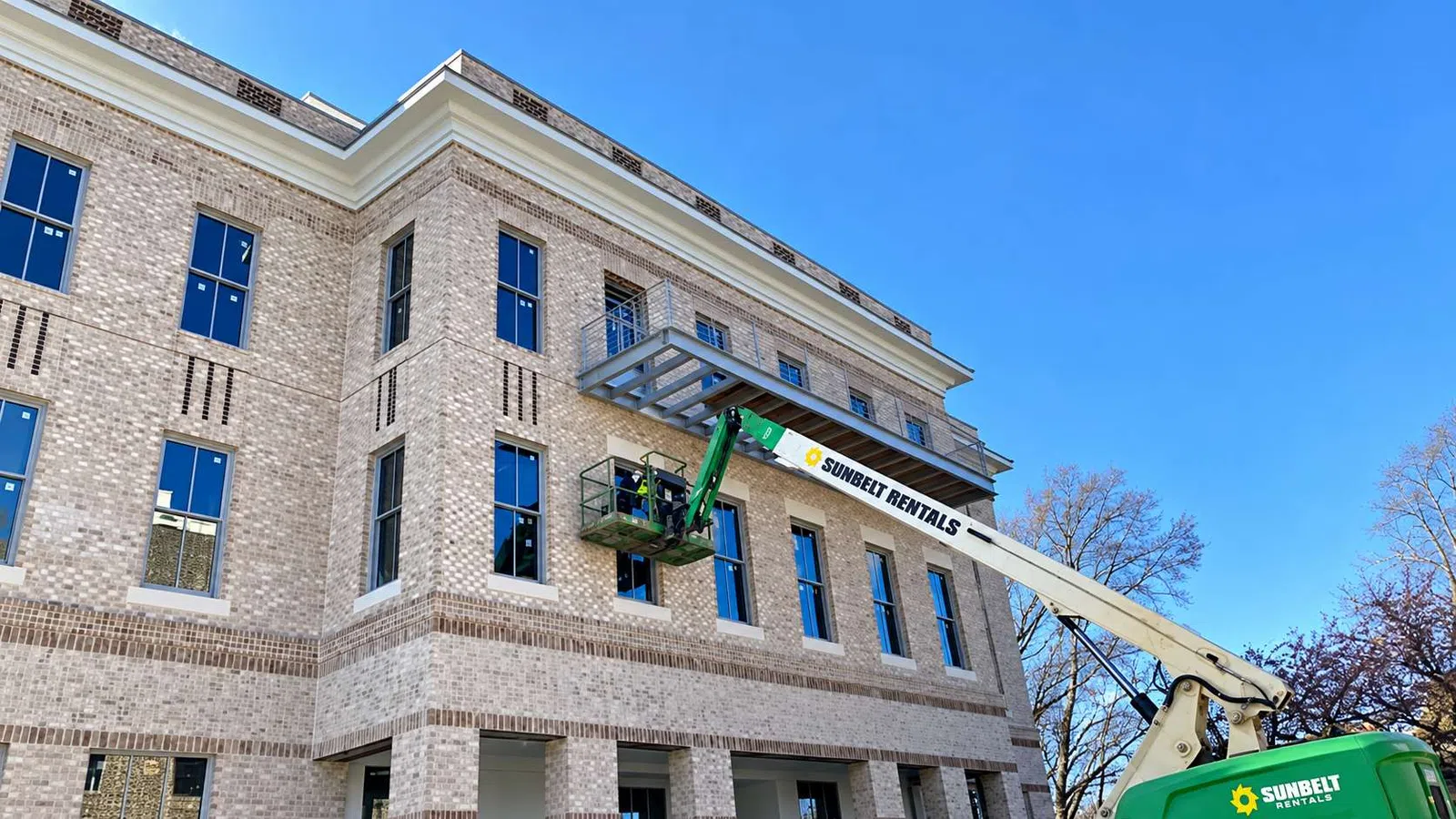 Construction workers using a Sunbelt Rentals lift to install a metal awning on a brick building under clear blue sky