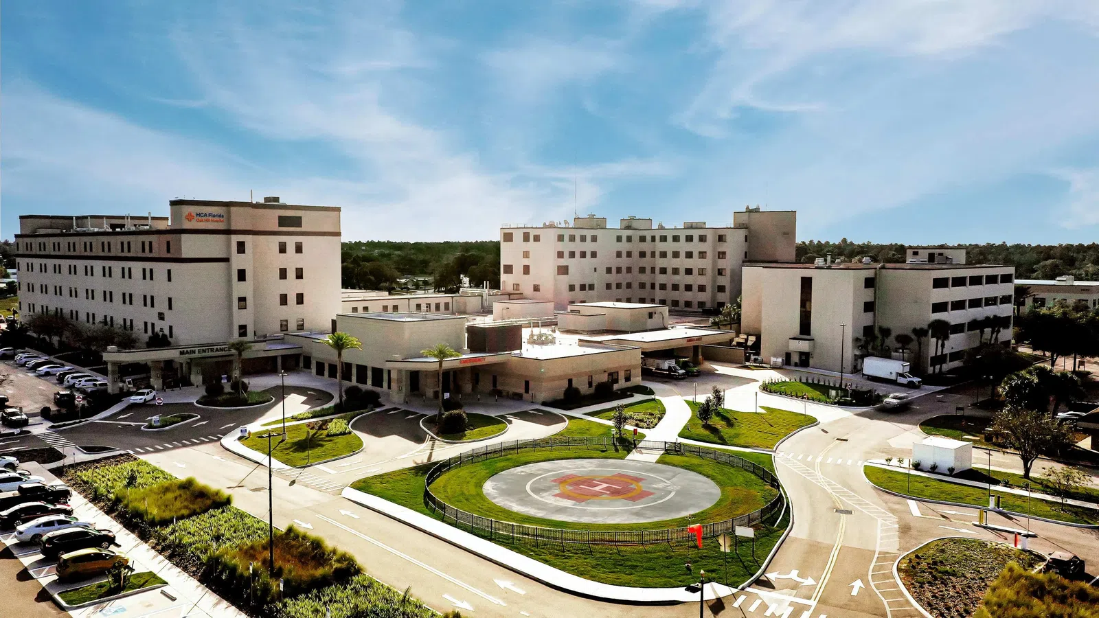 Aerial view of a large hospital complex with multiple buildings, parking lots, and a helipad in the center.