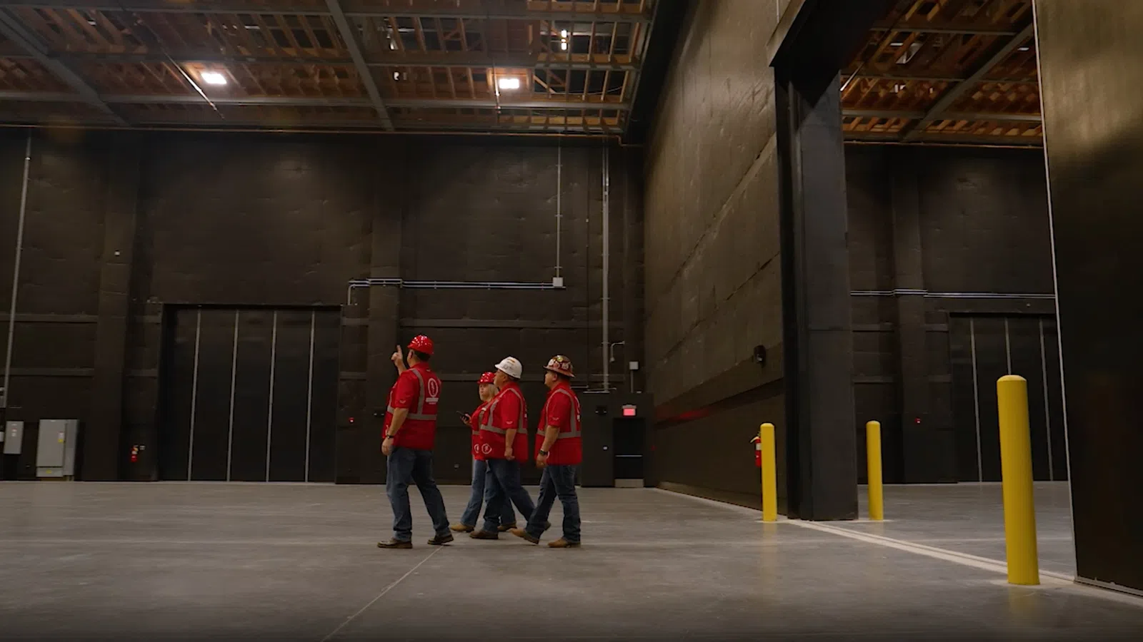 Four construction workers in red shirts and helmets inspect a large empty warehouse with concrete floors and high ceilings.
