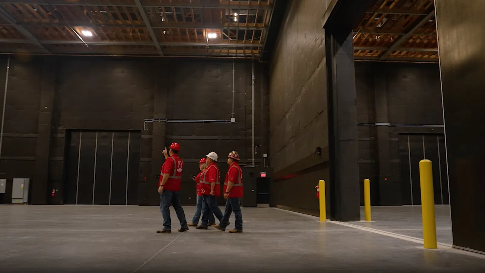 Four construction workers in red shirts and helmets walking inside a large empty industrial warehouse.