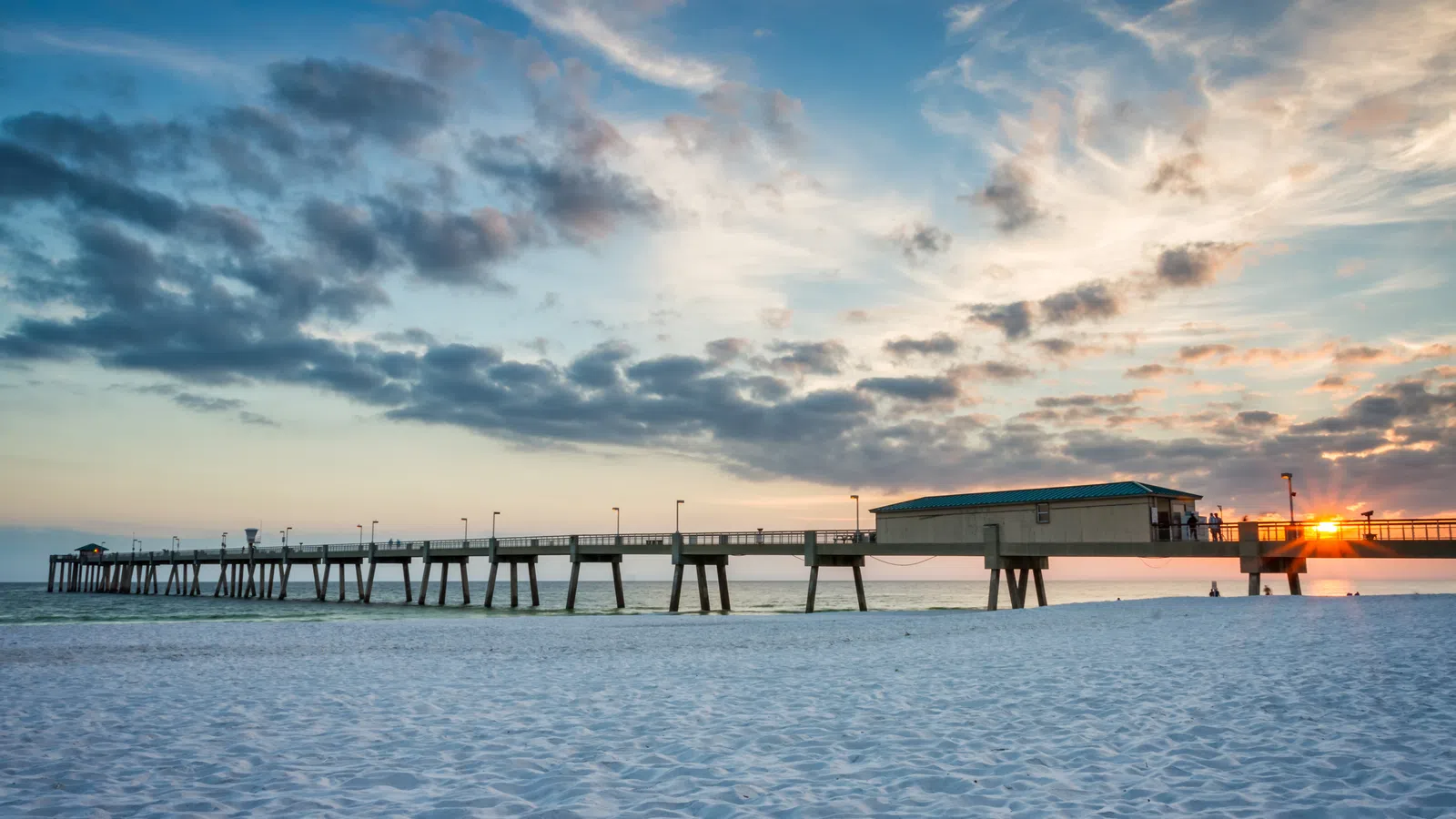 Long pier stretching over calm ocean waters at sunset under a partly cloudy sky over white sandy beach.