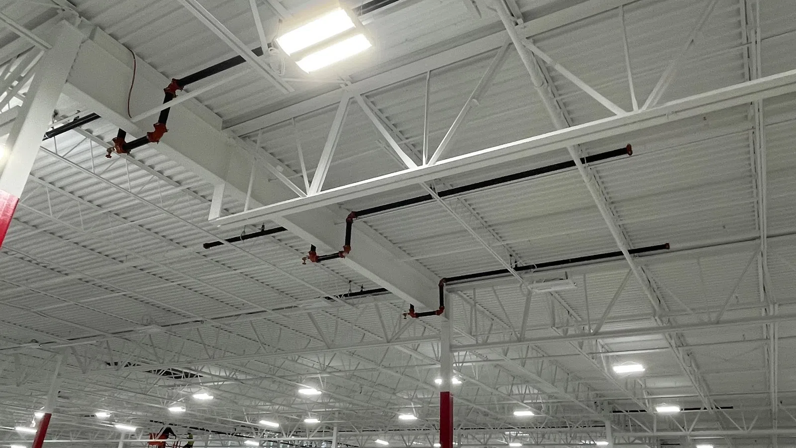 Interior view of a spacious industrial warehouse ceiling with white beams, pipes, and bright LED lights