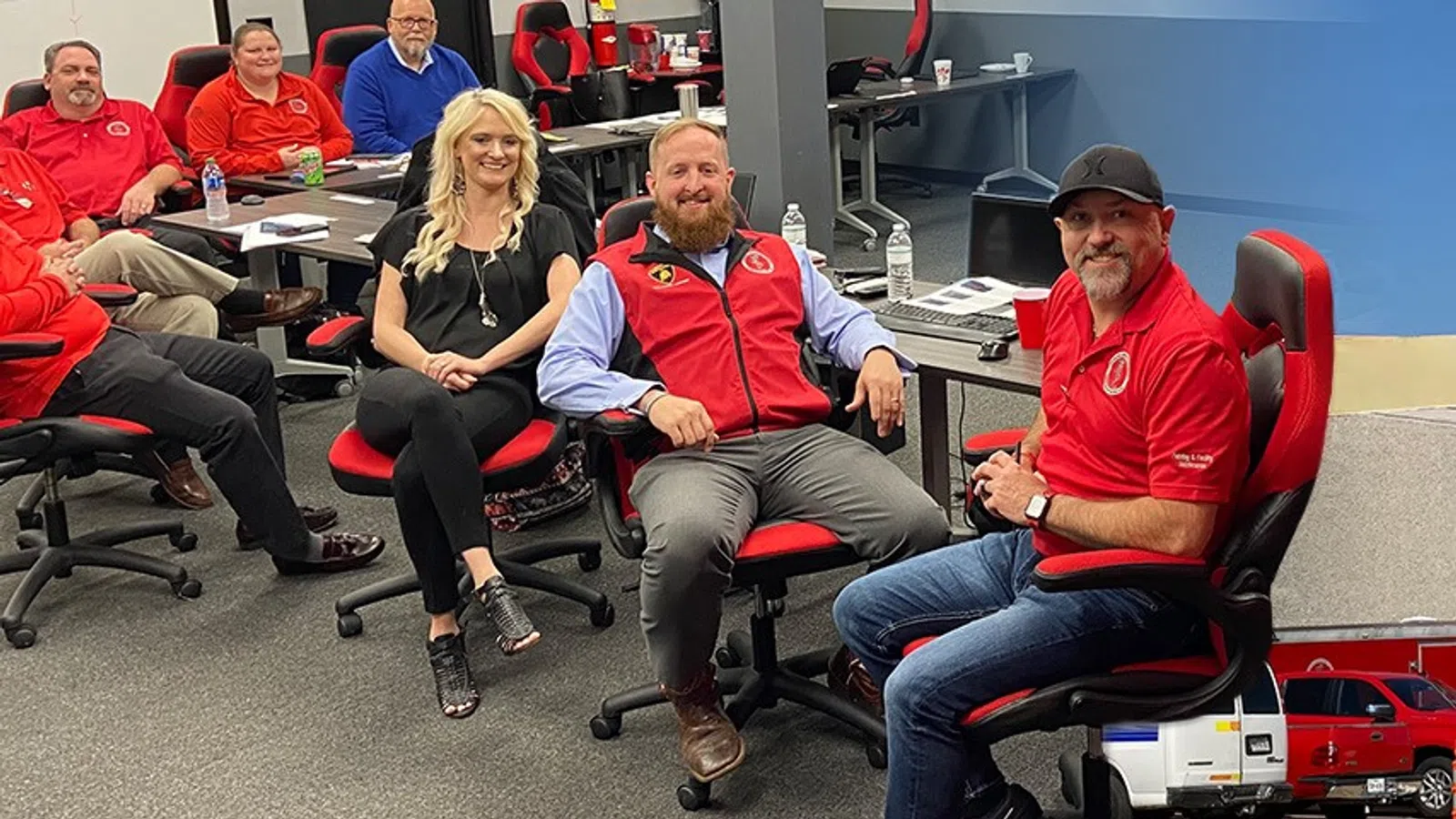 Group of professionals in red and black attire seated on office chairs in a modern meeting room.